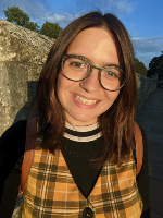 Brunette person with glasses, a black jumper and a tartan yellow scarf smiling in front of the York city walls